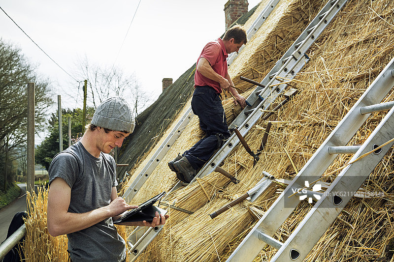 两位茅草匠在盖屋顶，一位使用平板电脑，另一位站在梯子上。图片素材