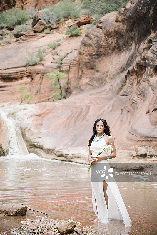 穿着白色长裙，站在河边，手持海芋的棕色长发年轻女人图片素材