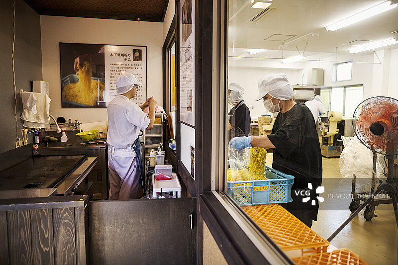 从为面馆提供服务的商业厨房看到的面条厂生产单元的景象，人们在工作，一位厨师在炉子旁。图片素材