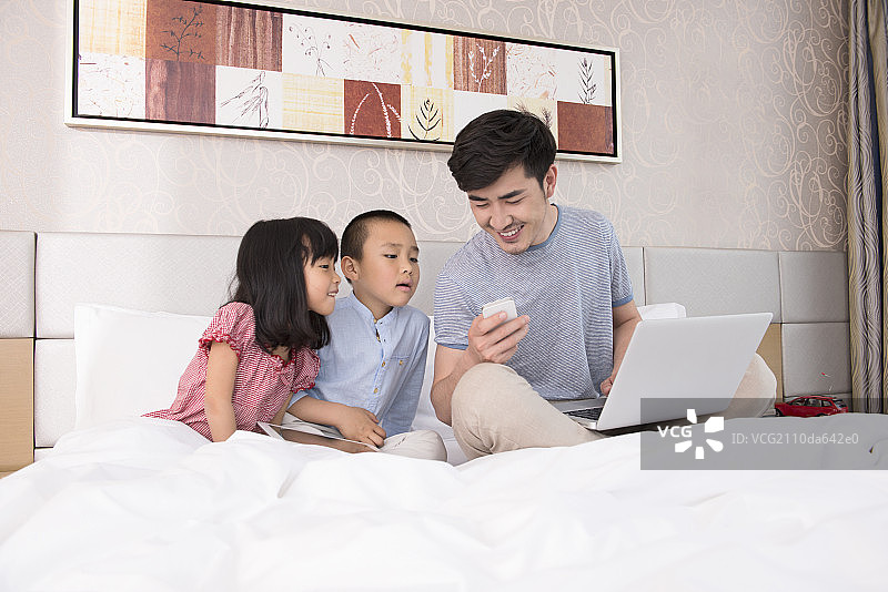 Father working on laptop in bed with children图片素材