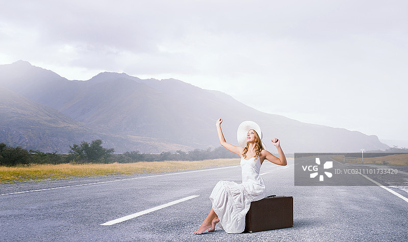 轻装旅行：身穿白色长裙、头戴帽子的女人坐在柏油路上的行李上图片素材