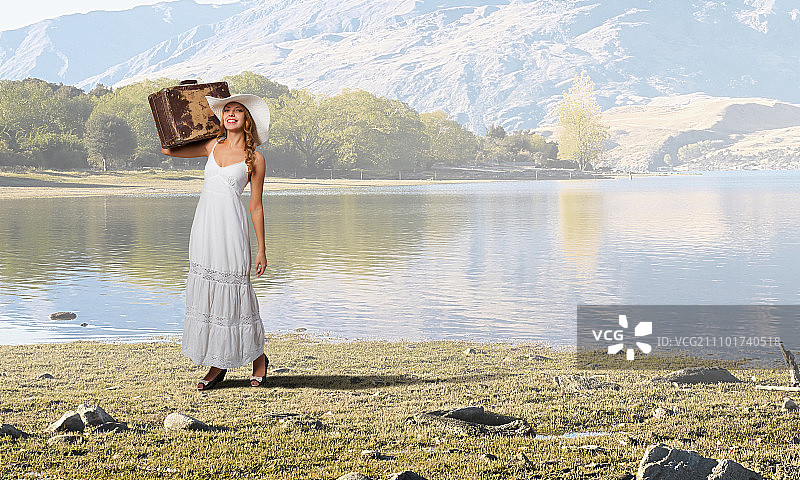 轻装旅行的女人：乡间道路上穿白色长裙、戴帽子的女士与手提箱图片素材