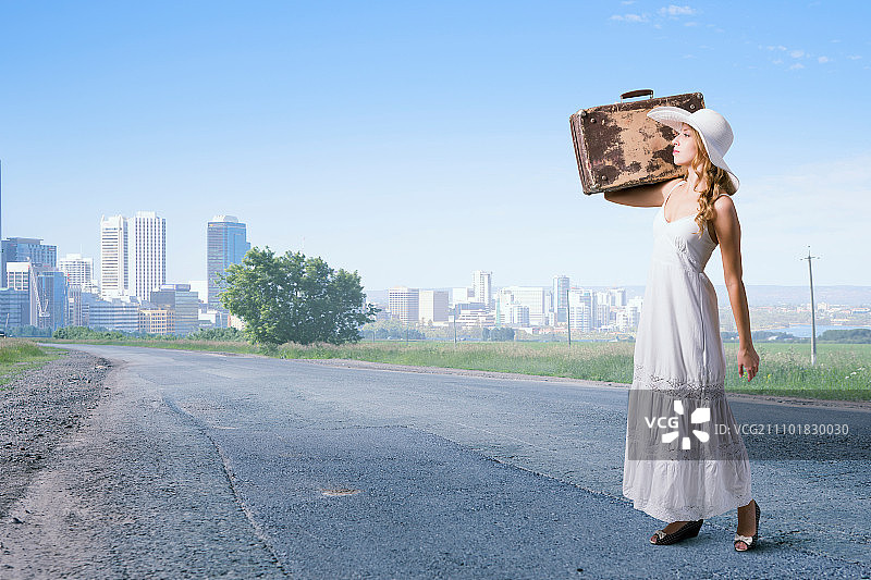 她正在轻装旅行：身穿白色长裙、头戴帽子的女人在沥青路上拉着行李箱图片素材