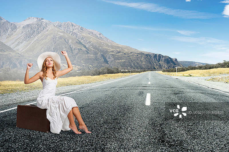 轻装旅行：身穿白色长裙、头戴帽子的女人坐在柏油路上的行李上图片素材