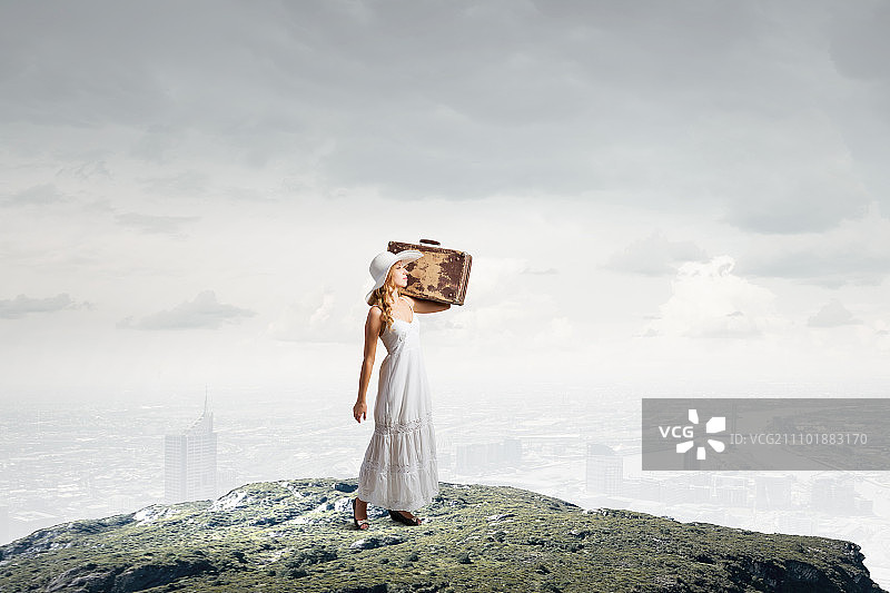 轻装旅行的女人：乡间道路上穿白色长裙、戴帽子的女士与手提箱图片素材