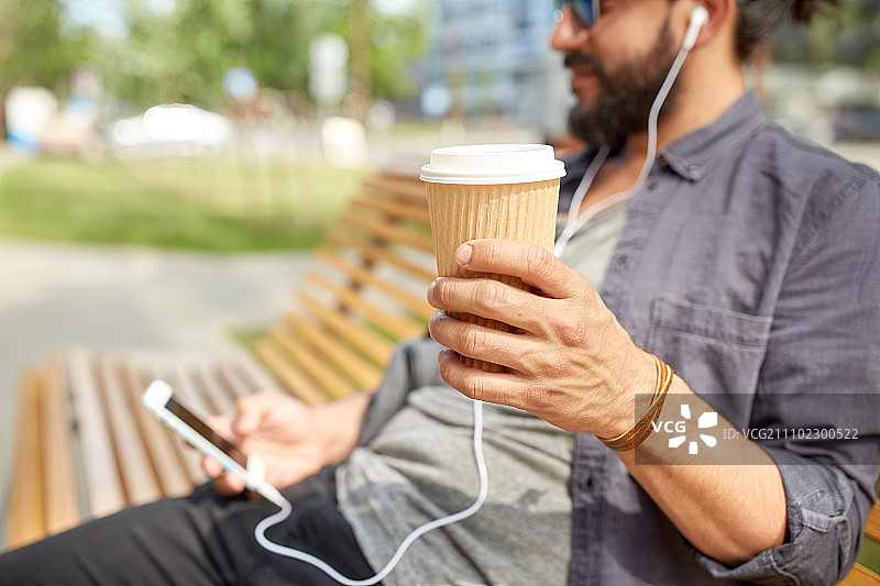 人物、饮品、技术、休闲和生活方式：戴着耳机和智能手机的男子在城市街道长椅上用一次性纸杯喝咖啡的特写图片素材