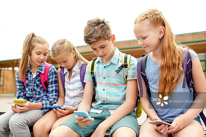小学教育，友谊，童年，科技和人们的概念 - 一群快乐的小学生带着智能手机和背包坐在户外长椅上图片素材