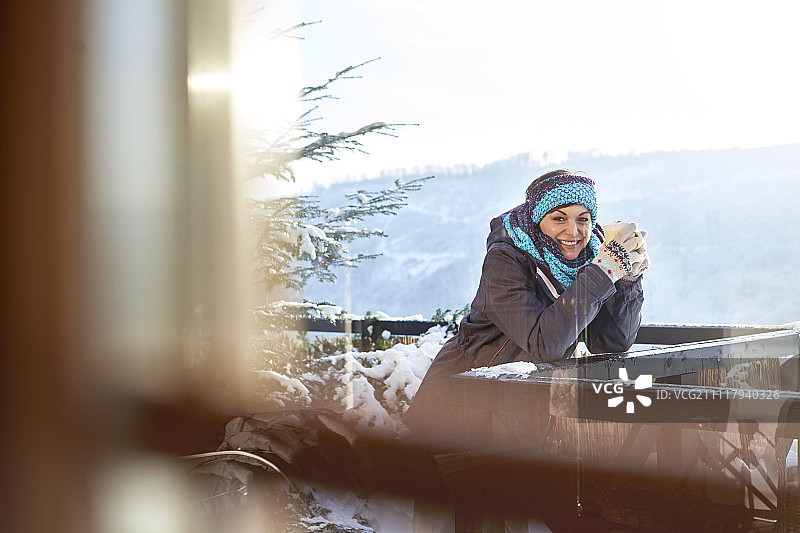 在阳光明媚的小屋阳台上喝热可可的微笑女滑雪者肖像图片素材