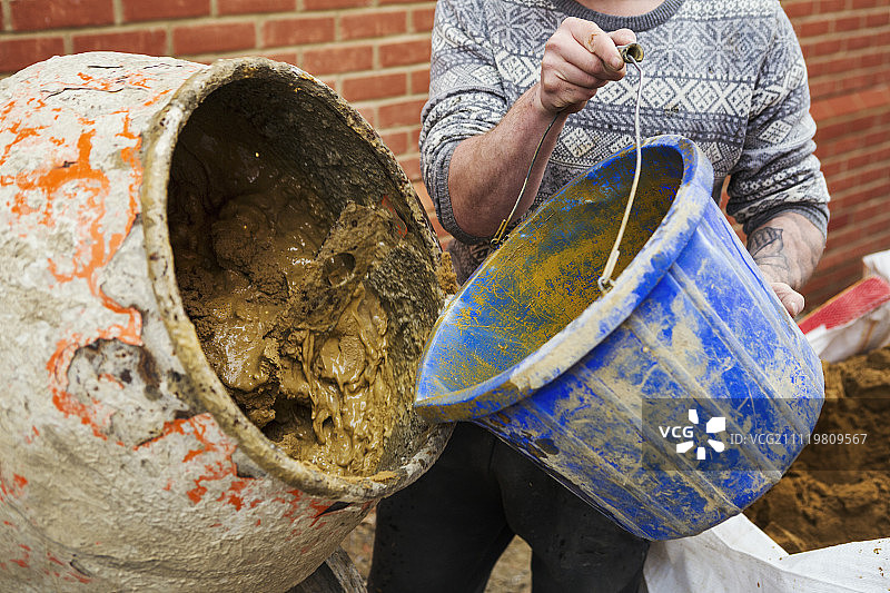 站在水泥搅拌机旁的建筑工人，砖匠拿着一个蓝色桶图片素材