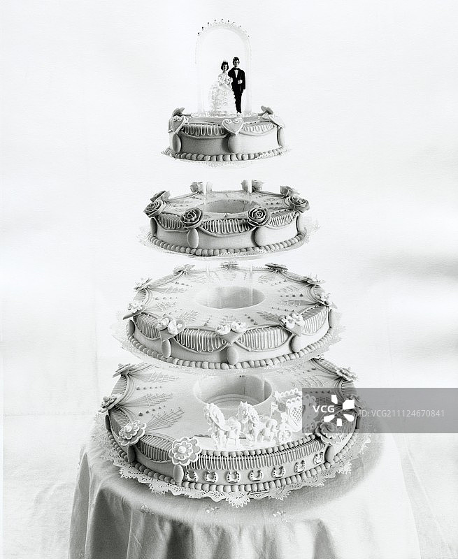 顶部带有新娘夫妇小雕像的四层白色婚礼蛋糕，黑白色图片素材