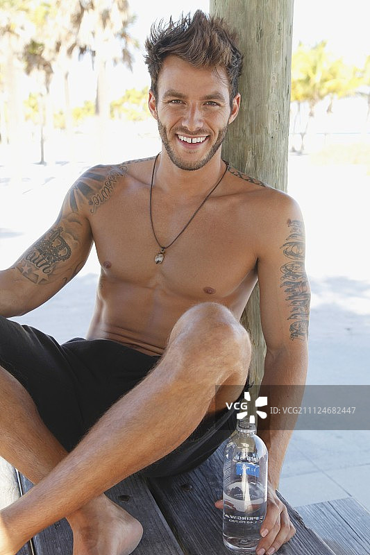 坐在树下穿着短裤拿着一瓶水的年轻纹身男子图片素材
