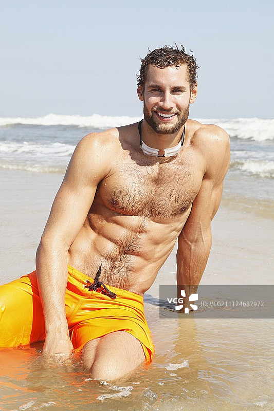 穿着黄色百慕大短裤坐在海滩浅水区的健壮年轻人图片素材