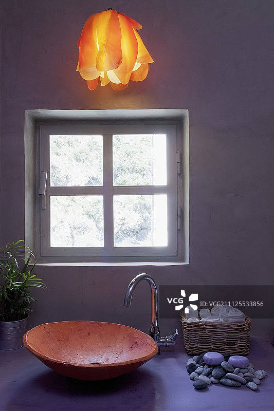 带有陶瓷洗手盆的氛围浴室和暖橙色，悬挂在暴露的混凝土墙前淡紫色梳妆台上方的灯图片素材