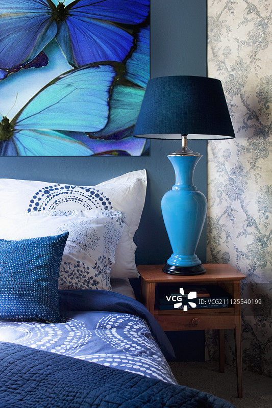 蓝色调的毯子、枕头和靠垫，蝴蝶图案上方；小柜子上放着蓝色床头灯图片素材