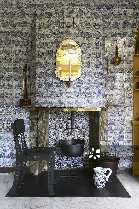 悬挂在开放式壁炉中的烹饪锅，整个烟囱胸墙和墙壁都铺有代尔夫特瓷砖；前景是带有穿孔结构的后现代金属椅子图片素材