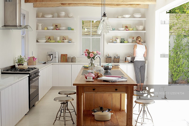 明亮、阳光明媚的厨房中独立的实木岛台，背景中一名妇女在厨房柜台旁图片素材