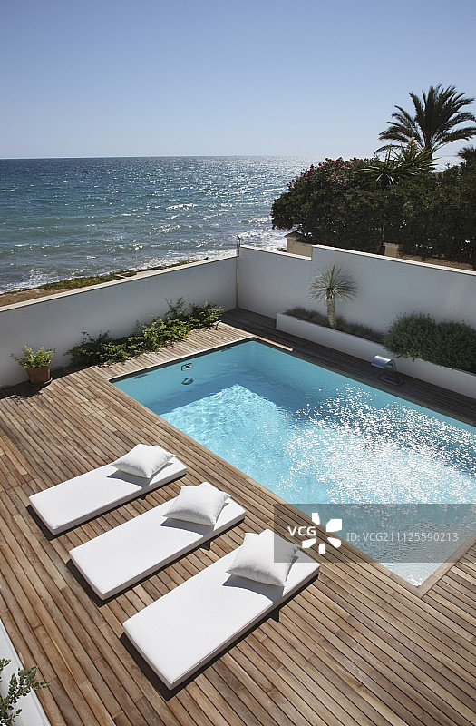 浅色蒲团日光躺椅与配套枕头在木制甲板上，泳池背景为海洋图片素材