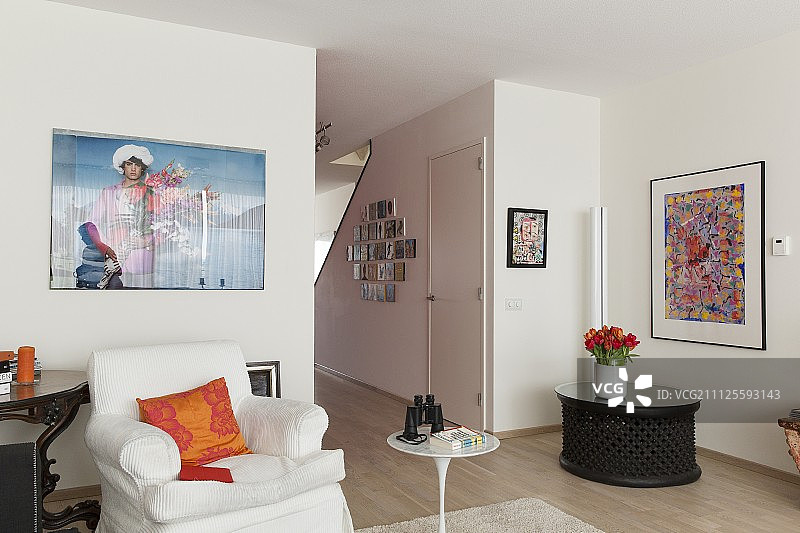 带橙色靠垫的白色扶手椅、经典郁金香边桌、低矮格子桌和墙上的艺术品图片素材