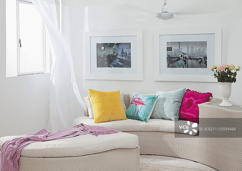 弯曲的浅色柳条沙发和脚凳，搭配彩色靠垫，位于墙上 framed 图片下方的角落图片素材