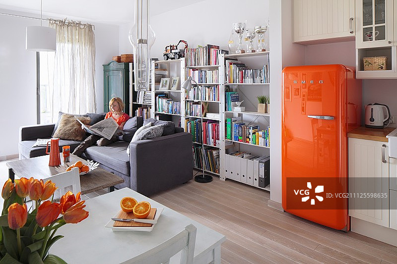 带橙色复古冰箱的开放式起居区；女人坐在书柜前的沙发上图片素材