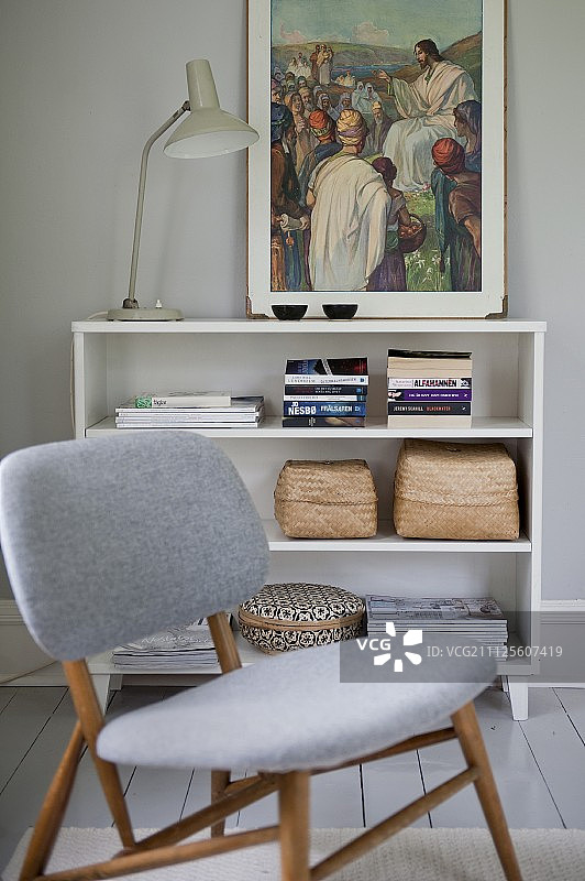 带有浅灰色软垫的椅子，位于复古台灯和半高搁架单元上的宗教绘画前图片素材