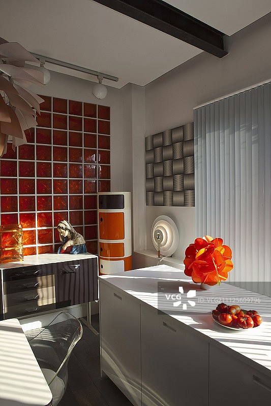 白色厨房台面上窗户下的橙色台灯和红色玻璃墙前的复古餐具柜上的圣母像图片素材