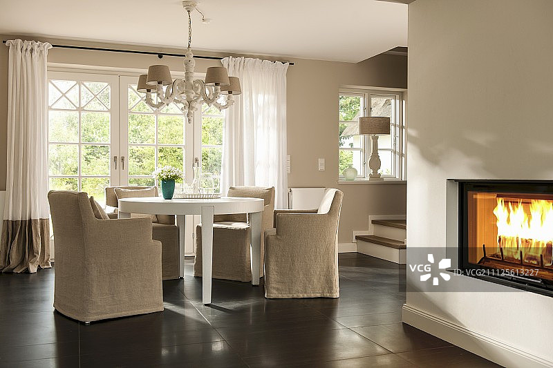 吊灯下白色圆桌旁的淡色宽松椅子，一侧壁炉里有火图片素材