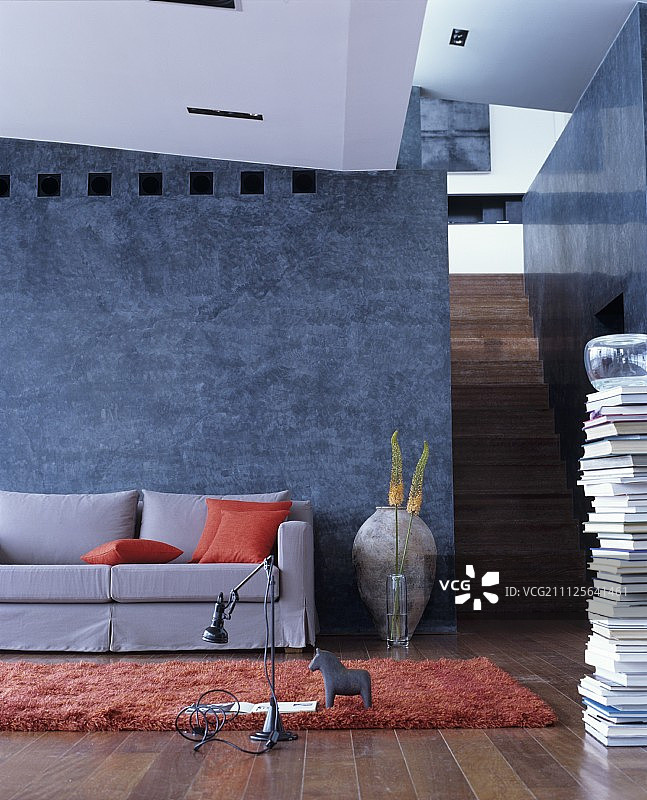 灰色的宽松沙发，带有橙色靠垫，背景是灰蓝色灰泥墙，一侧堆放着书籍和楼梯图片素材