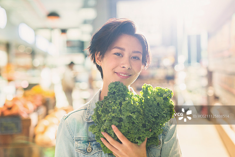 在杂货店市场里拿着新鲜羽衣甘蓝微笑的年轻女子肖像图片素材