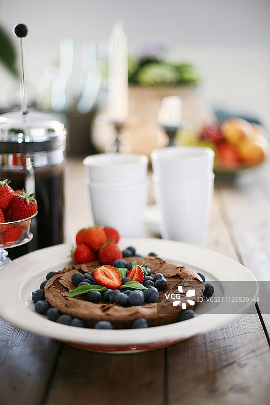 盘子上放着新鲜蓝莓和草莓的巧克力蛋糕，背景是白色杯子和咖啡壶图片素材