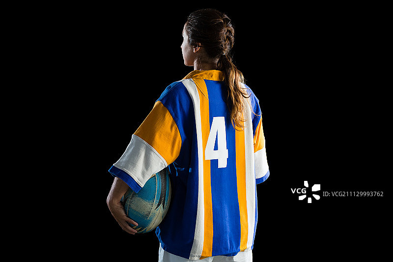 女运动员手持橄榄球，站在黑色背景前的背影图片素材