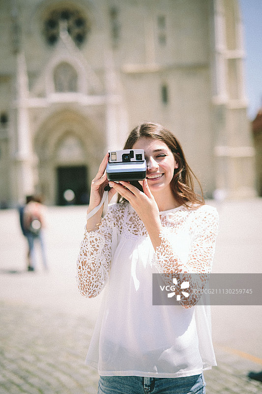 年轻女子用宝丽来相机在模糊的教堂前拍照图片素材