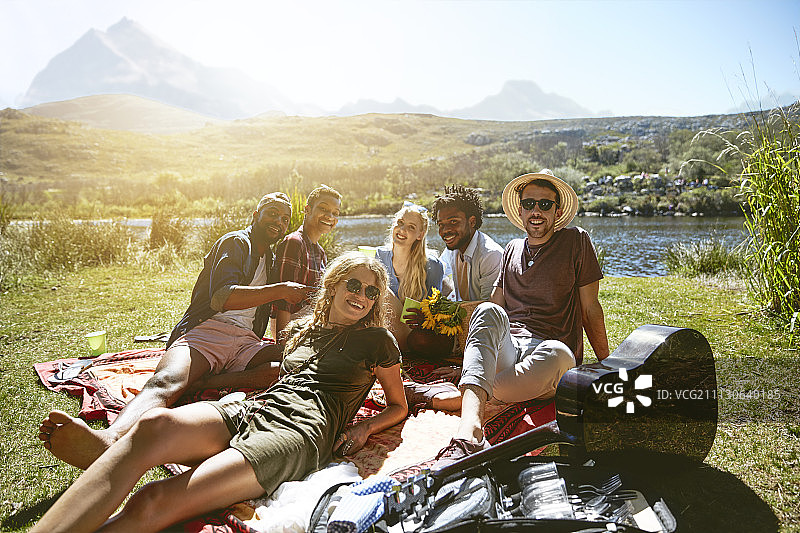 在阳光明媚的夏日河边享受野餐的微笑年轻朋友肖像图片素材