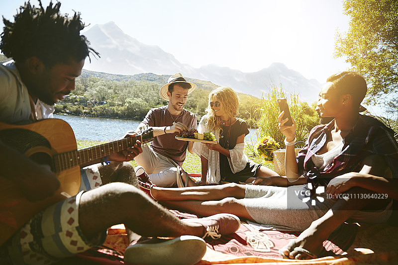 年轻的朋友们在阳光明媚的夏日河边闲逛、弹吉他、享受野餐图片素材