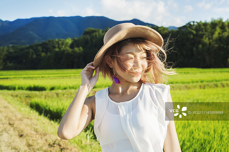 年轻女子在稻田和山景中，风中拿着帽子图片素材