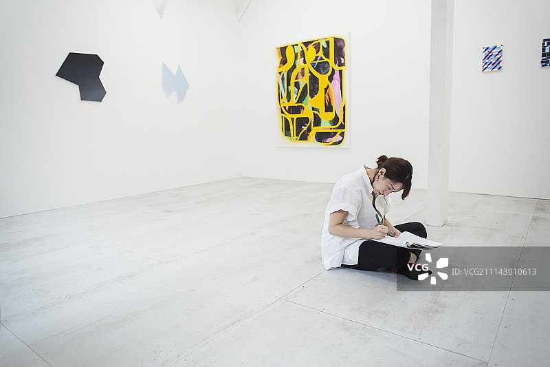 在艺术馆地板上坐着，手拿纸笔，身穿白色衬衫的黑发女子图片素材