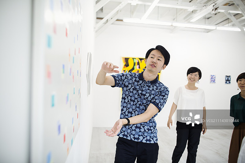 穿着蓝色衬衫的男人站在艺术馆白色墙壁上的现代绘画旁，背景中有两位女士图片素材