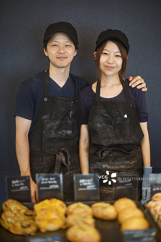 微笑的男人和女人戴着棒球帽和围裙站在面包店里，托盘里放着新鲜出炉的商品图片素材