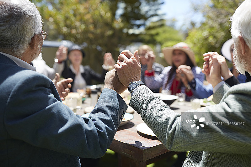 活跃的老年朋友手牵着手在阳光明媚的花园派对桌旁祈祷图片素材