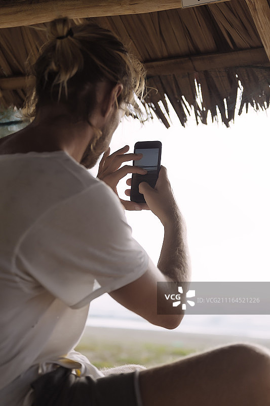 男子坐在印度尼西亚苏门答腊亚齐海滩的草棚下，用智能手机拍摄大海图片素材