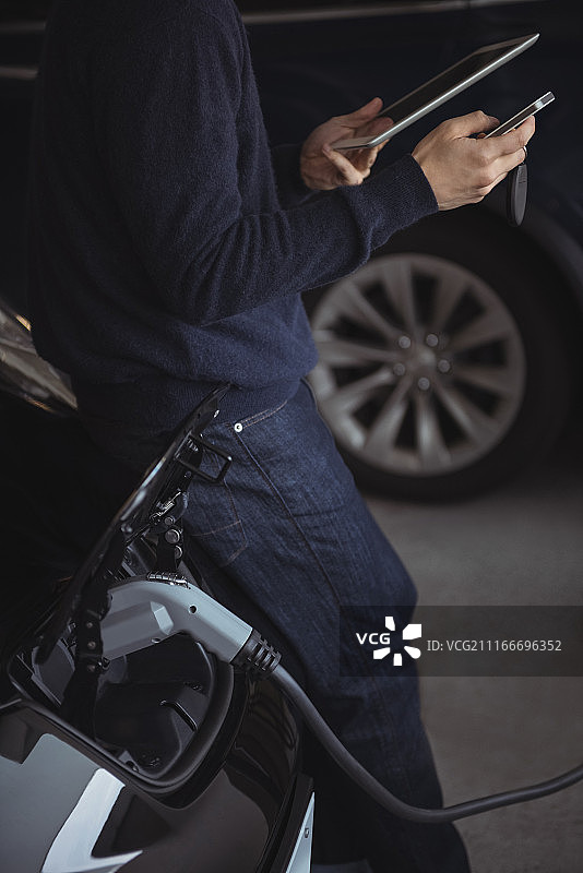 男子在给电动汽车充电时使用平板电脑和手机的腰部特写图片素材
