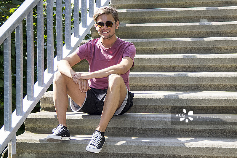 戴太阳镜、穿T恤和短裤坐在台阶上的英俊男人肖像图片素材
