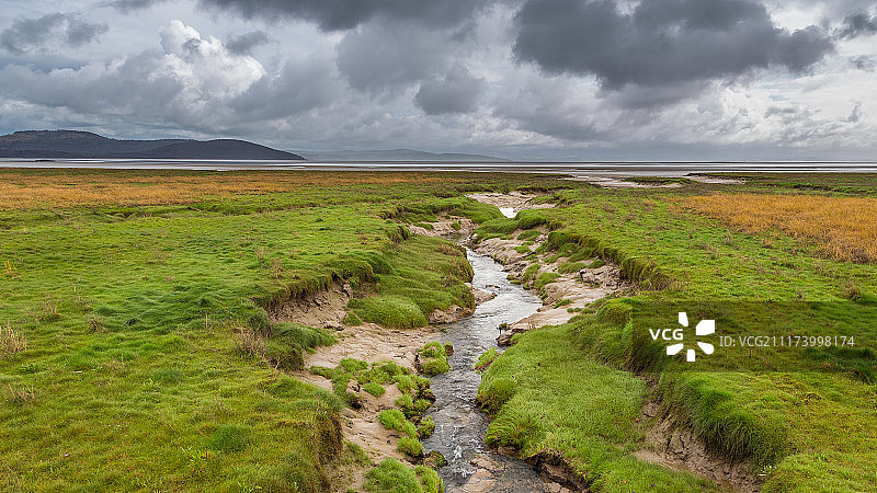 阴沉天空下，溪流汇入大海的草原图片素材