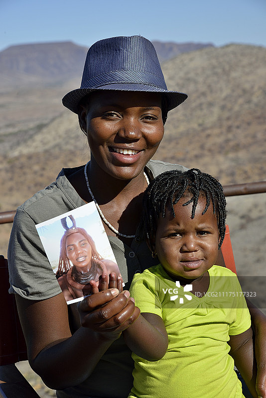 现代辛巴族妇女抱着孩子，背景是她自己穿着传统服装的照片，奥鲁佩姆贝，卡科兰，库内内，纳米比亚，非洲图片素材