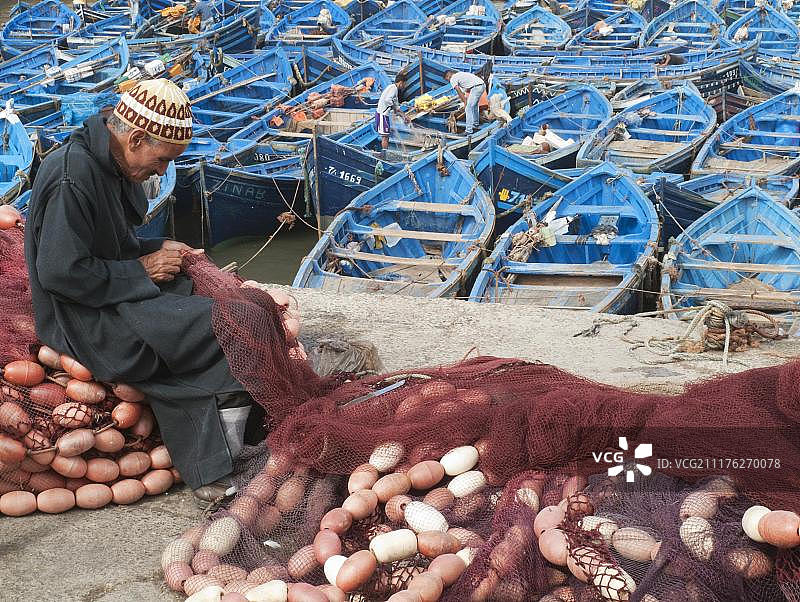 男人在索维拉渔港修理渔网，背后是蓝色渔船，摩洛哥，非洲图片素材