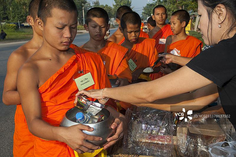 清晨的化缘，来自寺院学校的年轻佛教僧侣手持钵，接受一位年轻女子的食物或供品，泰国北部素可泰府，亚洲图片素材