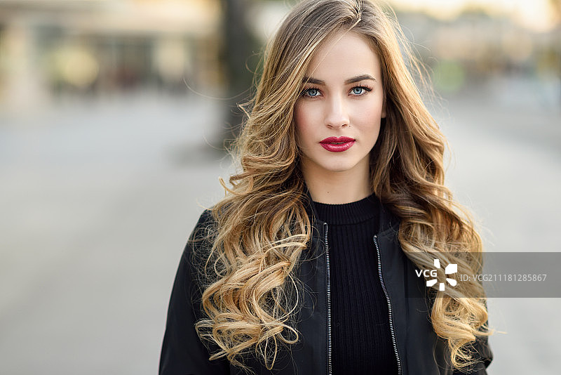 在户外穿着黑色夹克的有着美丽蓝眼睛的年轻金发女孩特写。漂亮的俄罗斯女性，有着长长的波浪发型。城市背景中的女人。图片素材