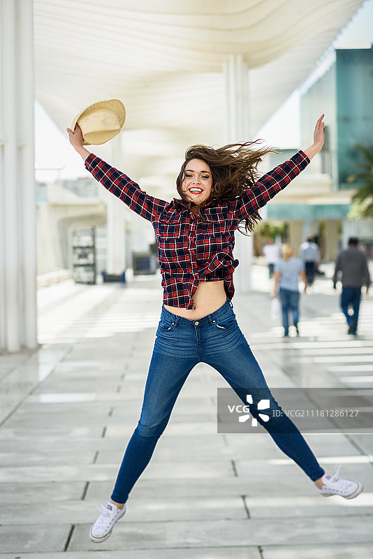 年轻女子戴着格子衬衫和遮阳帽，拥有美丽的蓝眼睛，在城市背景中跳跃图片素材