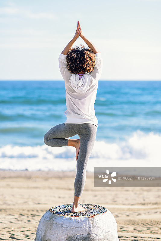 在海滩上做瑜伽的年轻黑人女子背影，穿着白色运动服的女子在树式中，背景是散焦的海景，非洲发型图片素材
