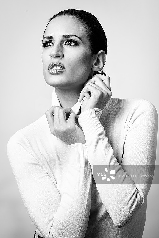 年轻的黑发女子穿着白色高领毛衣的影棚照。时尚模特黑白影棚照，白色背景图片素材